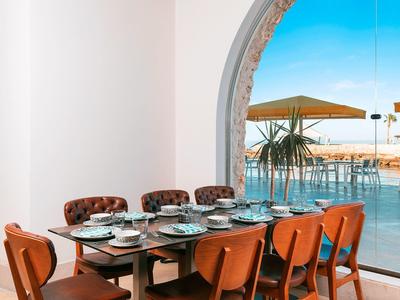 Elegant dining table with set plates and chairs, overlooking a terrace and sea through a large arch window.