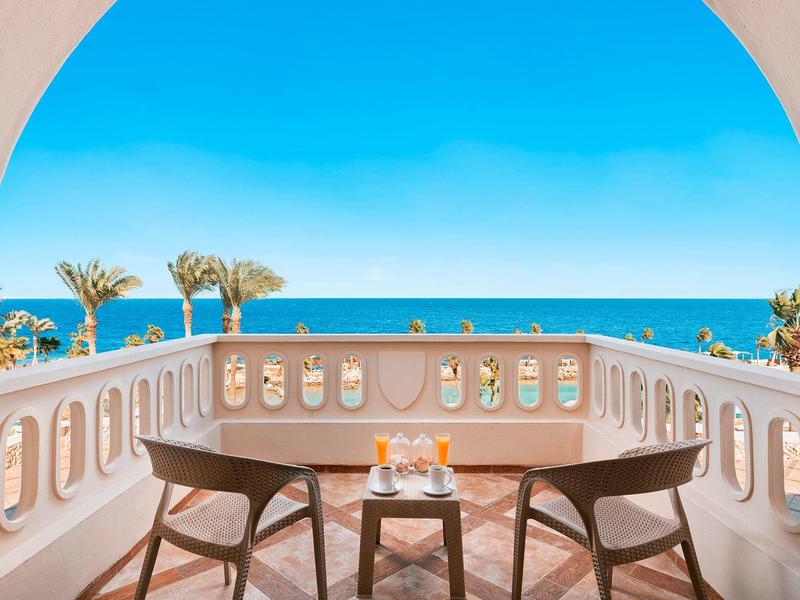 Balcony with two chairs and a table overlooking palm trees and the sea on a sunny day.