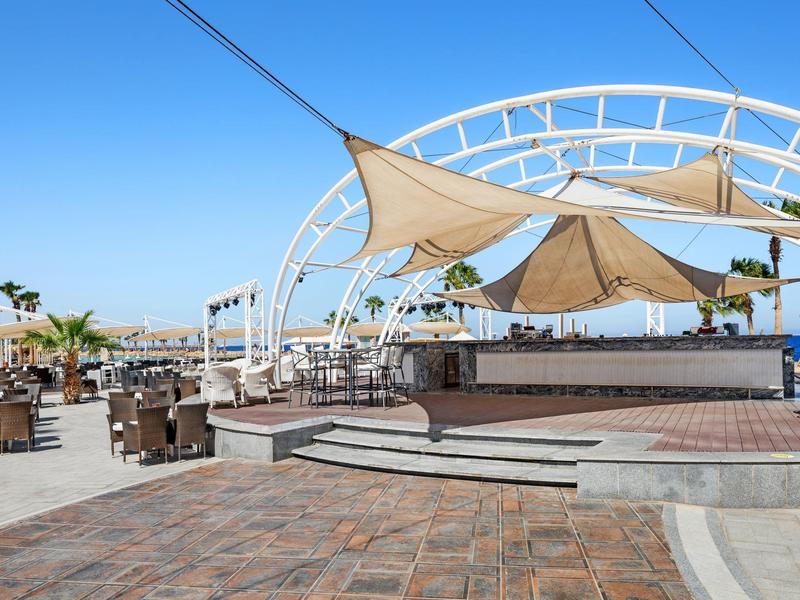 Sunlit outdoor seating area with chairs and tables under a shaded structure on a clear day.