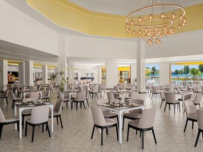 Przestronna jadalnia hotelowa w nowoczesnym stylu, beżowe krzesła, białe stoły i widok na ocean przez duże okna.