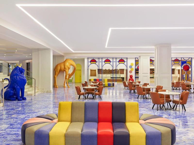 Nowoczesna lobby hotelowa z kolorowymi sofami, niebieskimi i żółtymi rzeźbami oraz stołami do jedzenia.