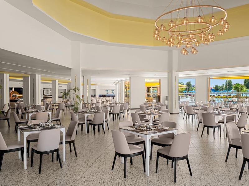 Przestronna jadalnia hotelowa w nowoczesnym stylu, beżowe krzesła, białe stoły i widok na ocean przez duże okna.