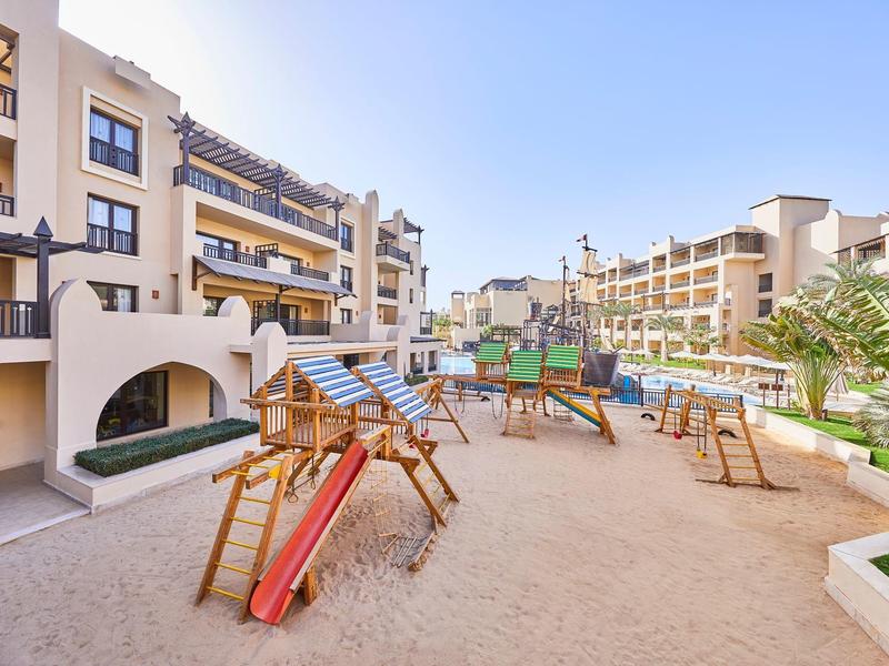 Outdoor playground with slides and climbing equipment in a sandy courtyard of a hotel.