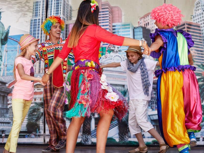 Children and adults in colorful costumes dancing outdoors with city buildings in the background.