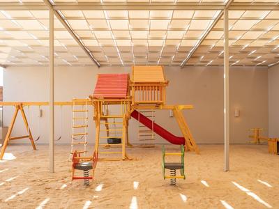 Indoor playground with wooden play structures, swings, slides, and sandy floor under a light grid ceiling.