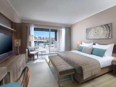 Modern hotel room with king bed, desk, TV, and balcony overlooking cityscape.