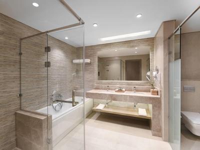 Modern hotel bathroom with glass shower, bathtub, large mirror, and beige tiles.