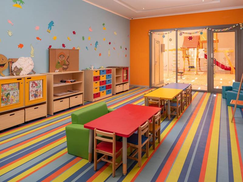 Kids' playroom with colorful striped carpet, small tables, chairs, shelves, and wall decorations.