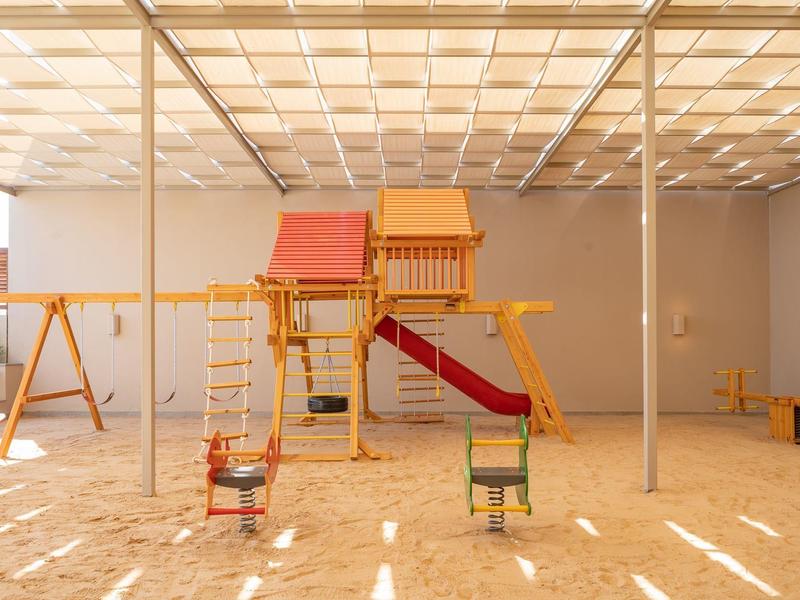Indoor playground with wooden play structures, swings, slides, and sandy floor under a light grid ceiling.