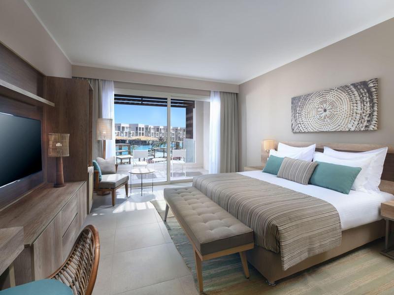 Modern hotel room with king bed, desk, TV, and balcony overlooking cityscape.