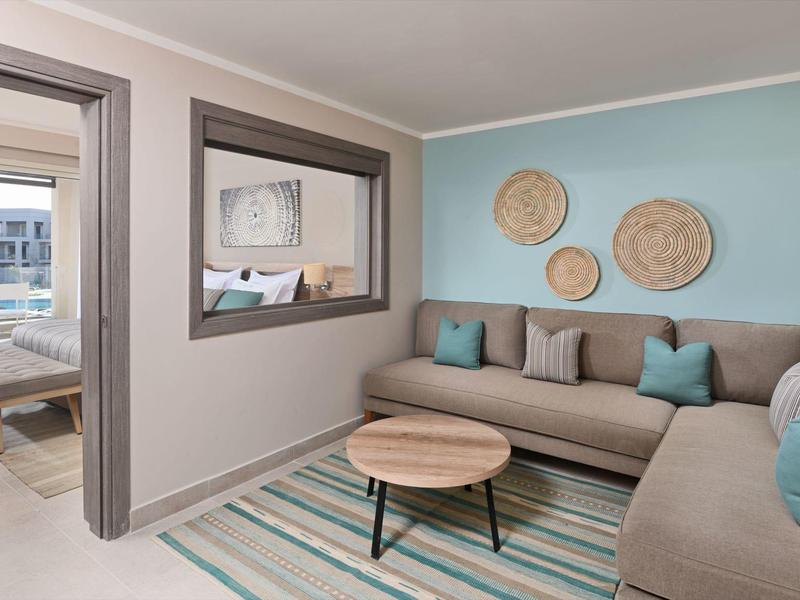 Modern hotel room with beige sectional sofa, round wooden table, and turquoise accents.