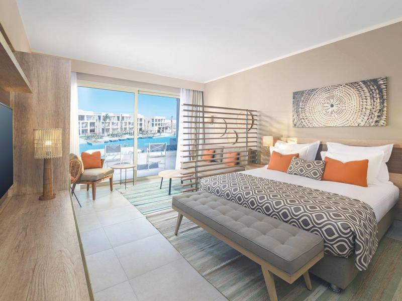 Bright hotel room with a large bed, balcony, and view of a marina under clear skies.