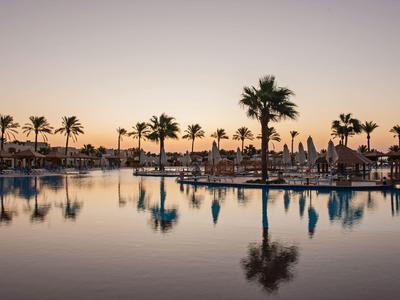 Une piscine calme au coucher du soleil avec des palmiers et des chaises longues en arrière-plan.