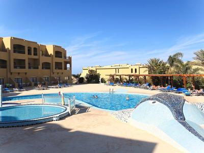 Großer Pool mit blauer Wasseroberfläche, umgeben von Sandsteinfarbenen Gebäuden und Palmen.