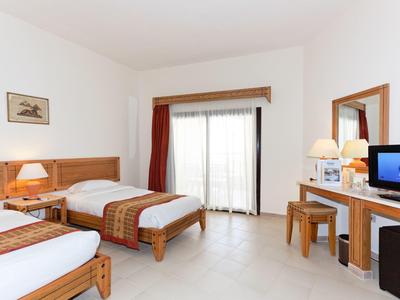 Helles Hotelzimmer mit zwei Einzelbetten, Holzmöbeln, Flachbildschirm und großem Fenster mit Vorhängen.