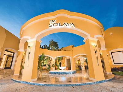 Hotel amarillo claro con arco y fuente bajo un cielo azul despejado.