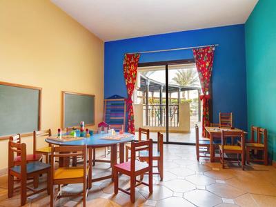 Sala de juegos colorida con mesas, sillas y grandes ventanas que dan al exterior.