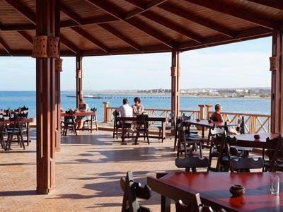 Überdachte Terrasse mit Holztischen und Stühlen, Blick auf das Meer und einen Hafen.