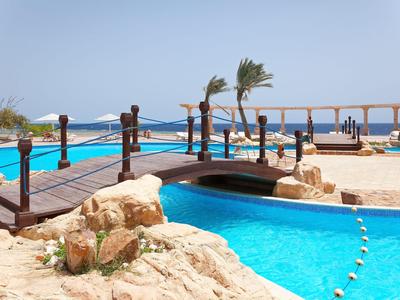 Poolbereich mit Brücke, Steinlandschaft und Blick auf das Meer unter klarem Himmel.