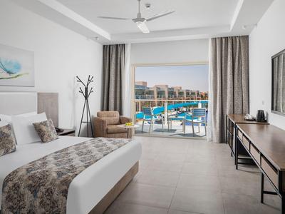 Moderne hotelkamer met kingsize bed, zithoek en balkon met uitzicht op zwembad en stad.