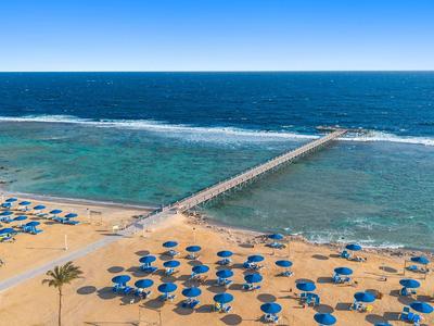 Strand met blauwe parasols en een lange houten pier die uitstrekt in de zee onder een heldere blauwe lucht.