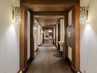 Long hotel corridor with wooden beams, wall lamps, and brown flooring leading to a distant seating area.