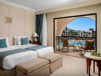 Hotelzimmer mit zwei Betten und Balkon mit Blick auf einen Pool und angrenzende Gebäude.