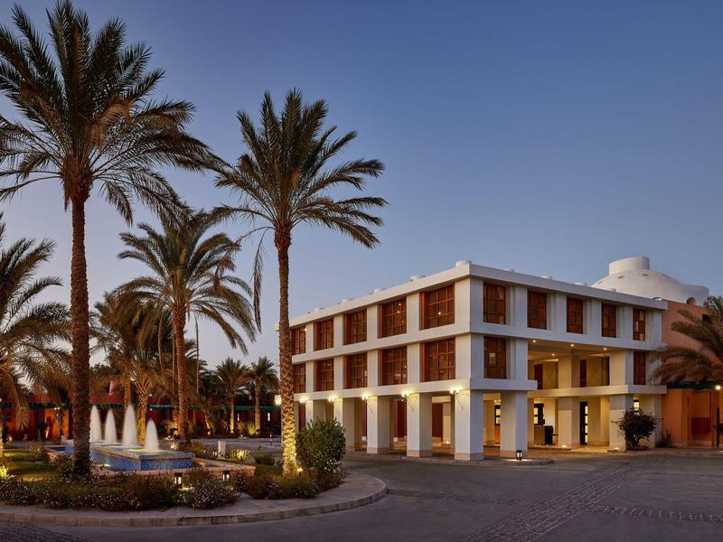 Hotel at dusk with palm trees and fountains in front, under a clear blue sky.