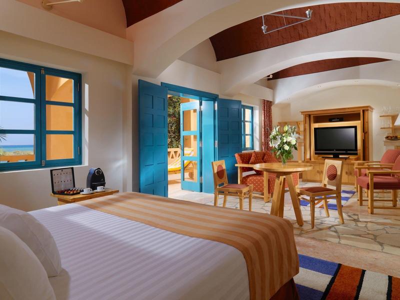 Cozy hotel room with a large bed, blue window frames, and a dining area with a TV.
