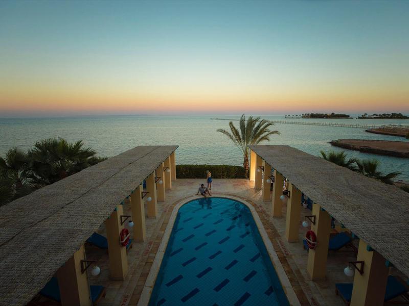 Sunset view over a hotel pool with palm trees and ocean in the background.