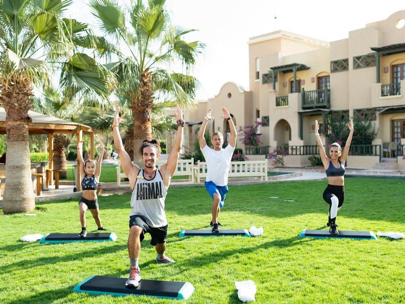 Grupo haciendo yoga en césped verde frente a hotel con palmeras y sombrillas.
