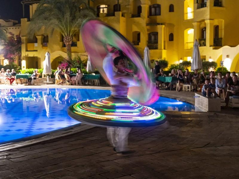 Timbalero iluminado con colores frente a una piscina con público en un hotel por la noche.