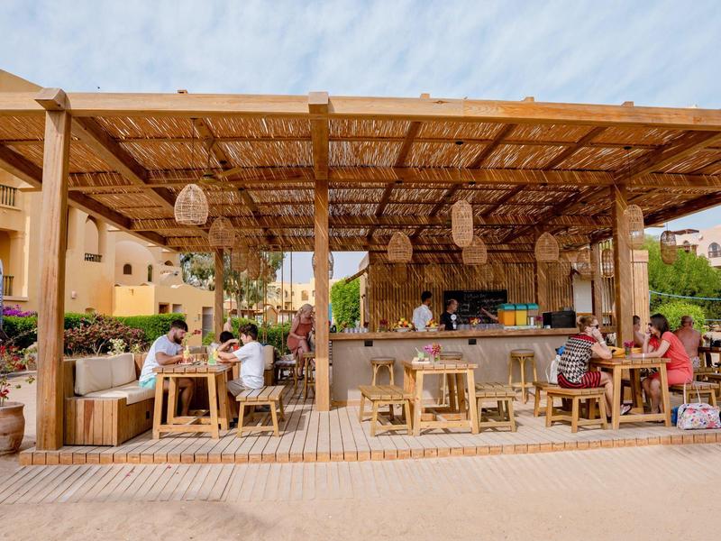 Bar abierta con techo de madera, clientes en mesas, asientos y ambiente mediterráneo.