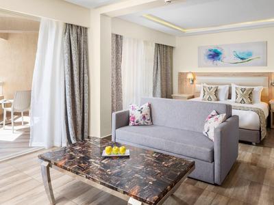 Lichte hotelkamer met bed, bank, marmeren salontafel en balkon met tafel en stoelen.