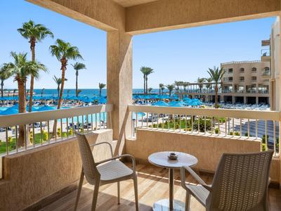 Balkon met stoelen en tafel met uitzicht op zwembad, palmbomen en hotel onder een heldere lucht.