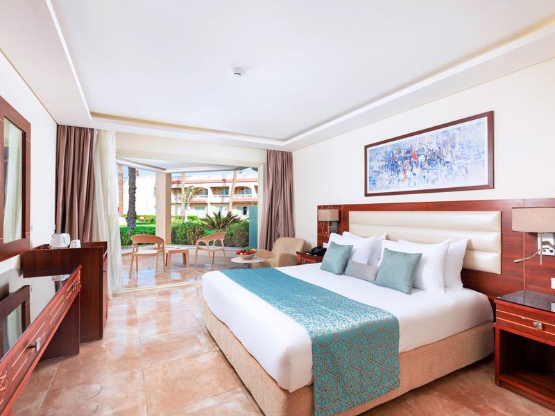 Ruime hotelkamer met groot bed, tv en zitgedeelte op het balkon zichtbaar door glazen deuren.