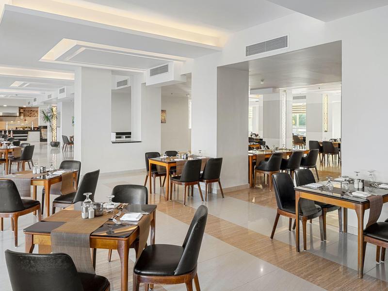 Ruime eetzaal van hotel met moderne houten tafels en zwarte stoelen, heldere witte muren en plafond.