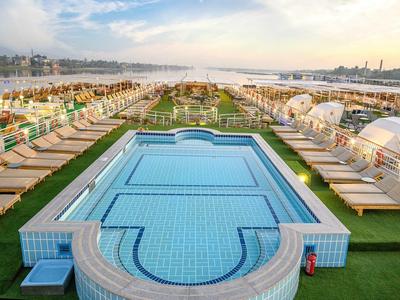 Großer Pool mit blauen Fliesen, umgeben von Liegen und Sonnenschirmen am Meer bei Sonnenuntergang.