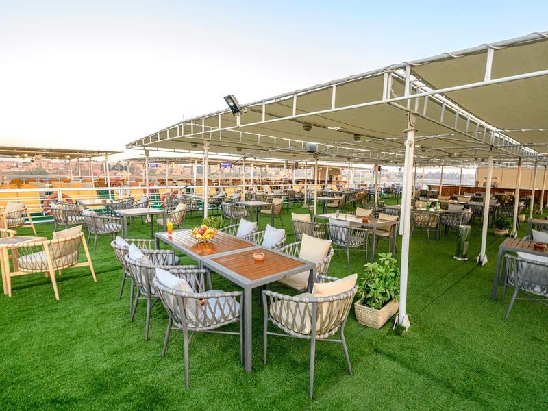 Buiten eetgedeelte van hotel met tafels, stoelen en schaduwdoek op kunstgras.