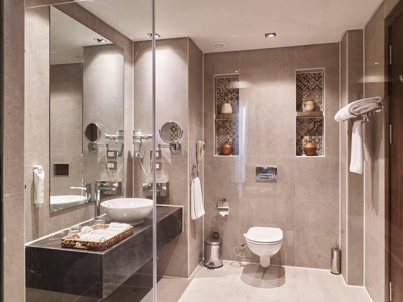 Baño moderno de hotel con lavabo, espejo, inodoro y ducha accesible en tonos neutros.