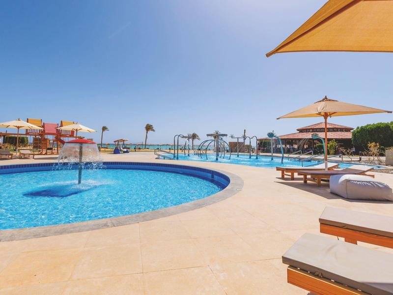 Piscina al aire libre con paraguas, camas y palmeras bajo un cielo claro y soleado.