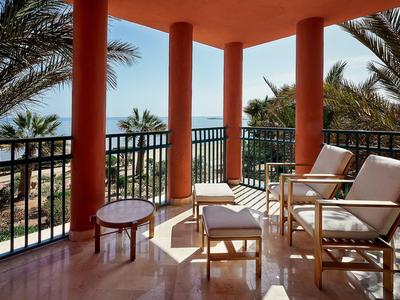 Balkon met stoelen en tafel, omgeven door rode zuilen en palmbomen.