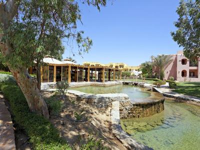 Hotel tuin met een vijver omgeven door bomen en gebouwen onder een heldere blauwe lucht.