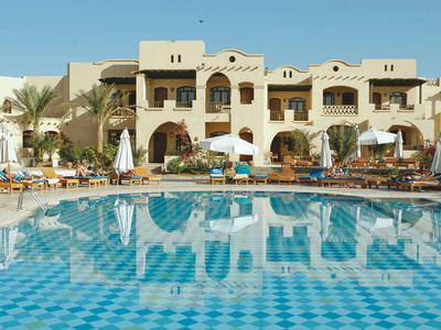 Hotel met groot buitenzwembad, ligstoelen, parasols en een tweeverdiepingen hoog beige gebouw.