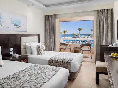 Hotel room with two beds, balcony overlooking the sea, chairs, and table.