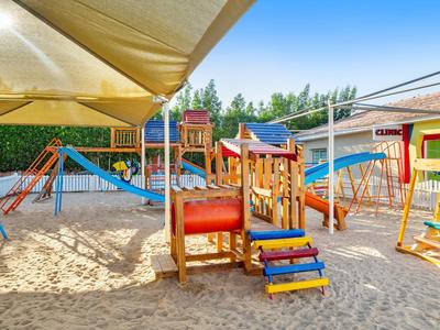 Parque infantil colorido con toboganes, columpios y estructuras de escalada sobre arena bajo toldos.