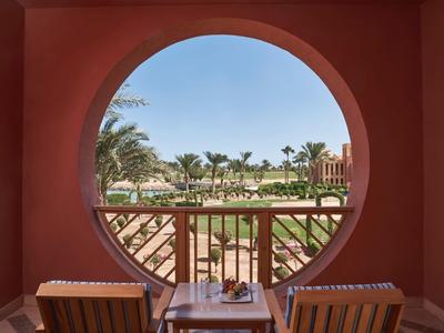 Výhled na tropickou zahradu přes kulaté okno s dvěma dřevěnými židlemi.