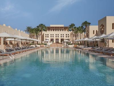 Zone piscine luxueuse avec chaises longues et parasols devant un grand bâtiment hôtelier sous un ciel bleu.