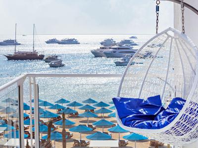 Silla colgante blanca con cojines azules en balcón con vista a la playa y barcos en el mar