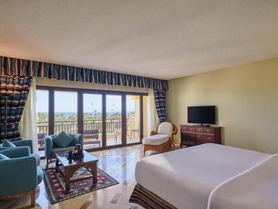 Helles Hotelzimmer mit großem Bett, Sitzbereich und Blick auf eine Terrasse und das Meer.